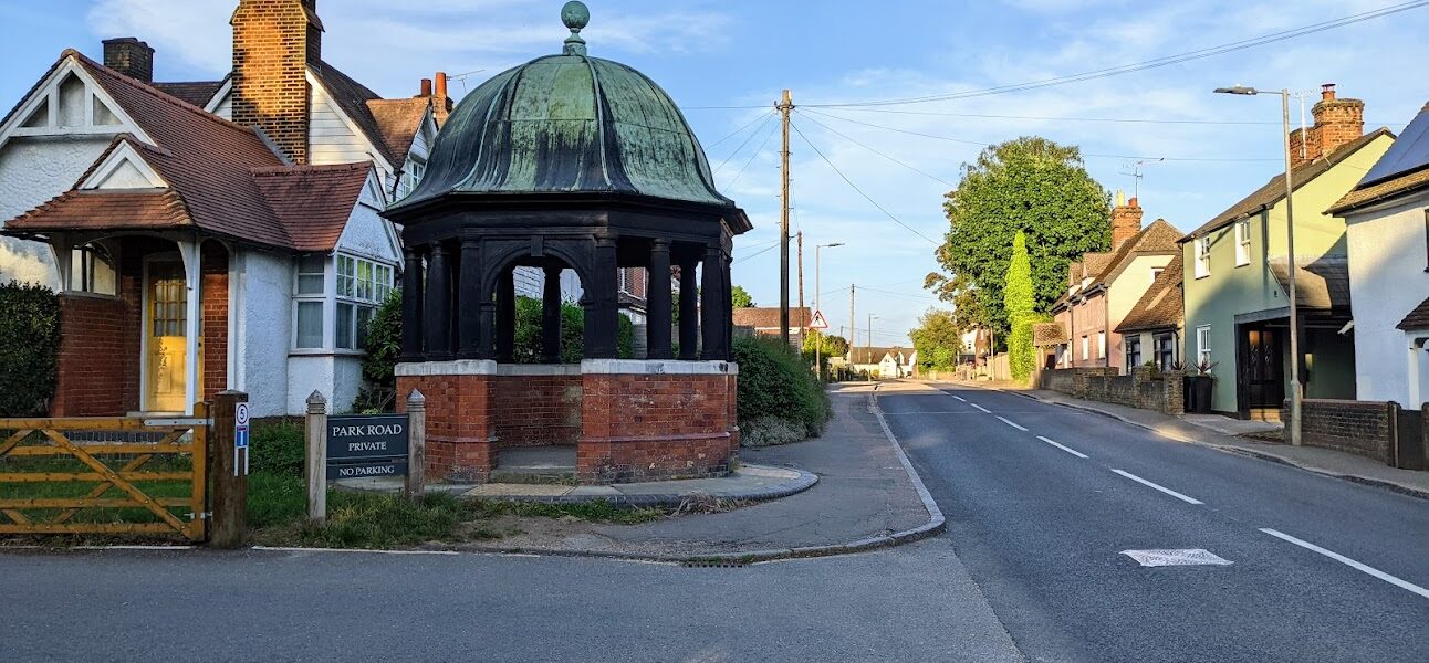 The Pump House and High Street in Elsenham. The Pump House is an octagonal structure with a red tiled wall, with wooden arches supporting a green domed roof.